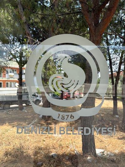 The image shows a window with a white logo and text. The logo is circular and features a stylized bird, possibly a rooster, surrounded by concentric circles. Below the logo, the text "DENİZLİ BELEDİYESİ" is visible, which translates to "Denizli Municipality" in English. The year "1876" is also part of the logo.
The scene outside the window is partially visible, showing trees with rough bark, dry grass and straw on the ground, and a building with orange and white facades in the background. A silver truck and a silver car are parked in front of the building. Reflections on the window include tree branches and foliage. The lighting suggests it is daytime with sunlight. The overall impression is that this is the exterior of a building in Denizli, Turkey, likely a municipal office or public facility, as indicated by the prominent logo.