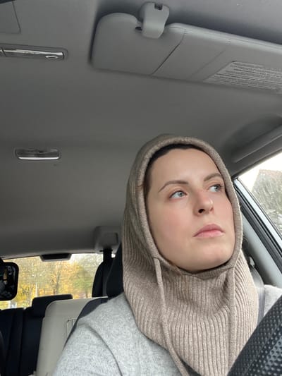 The image displays a female individual seated in a vehicle, located in Staudernheim, Germany. She occupies what appears to be the driver's position, with a black steering wheel partially visible in the lower right foreground. The individual possesses light skin, blue eyes, and wears a light grey long-sleeved garment underneath a ribbed, light beige knitted hood or balaclava, which features visible drawstrings. A dark seatbelt strap crosses her left shoulder and chest. Her gaze is directed upwards and to the right, away from the camera, maintaining a neutral facial expression. The vehicle's interior has a light grey ceiling, fitted with two rectangular overhead lights and a light grey sun visor on the passenger side. A rearview mirror is partially visible on the left, reflecting a portion of the car's cabin. In the rear seating area, a black car seat is visible, and behind it, a light-colored object resembling a pet carrier. Outside the vehicle, visible through the windows, are trees exhibiting yellow and orange autumn foliage, indicating the season. The lighting suggests an overcast sky, and a glimpse of a building with a reddish roof is visible through the passenger-side window.