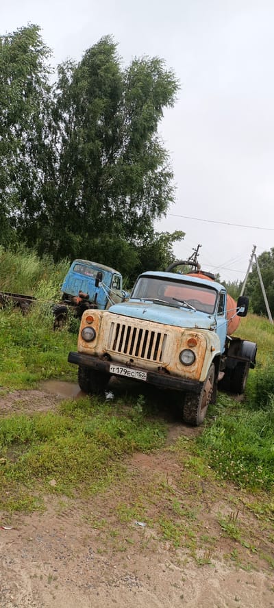 The image shows two dilapidated Soviet-era trucks parked in a grassy, muddy area with trees in the background. The truck in the foreground is light blue and white with a rusty grille and a license plate that reads "T 179EC 152 RU". It appears to be a tanker truck with a large orange tank visible behind the cab. The second truck, further in the background and partially obscured, is entirely blue and also appears to be in disrepair. The environment suggests an outdoor, possibly rural or abandoned location, with overcast skies and overgrown vegetation. There are no people visible in the image. The location is identified as Obzherikha, Russia.