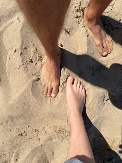 The image shows two pairs of bare feet standing on a sandy beach, likely in Lilaste, Latvia, given the context. The feet on the left belong to a person with tanned skin and visible veins, appearing to be male, with a tattoo on the ankle. The feet on the right belong to a person with paler skin, appearing to be female. Both sets of feet are partially sunk into the sand, creating shadows from an unseen light source. The environment is characterized by dry, light-colored sand with small debris and indentations. The image is taken from a top-down perspective, focusing on the feet and the immediate sandy surroundings.