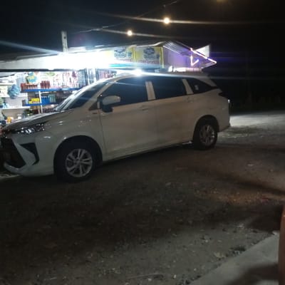 A white SUV is parked on a gravel lot at night in front of a small store. The store is illuminated by overhead lights and has shelves stocked with various items. A banner on the store reads "BELI 4 GRATIS 1" (Buy 4 Get 1 Free) in Indonesian. The time of day is night, and the weather appears to be clear. The location is Keudenibong, Indonesia. The scene appears to be ordinary, with no apparent emotions or specific events taking place beyond the store being open for business.
