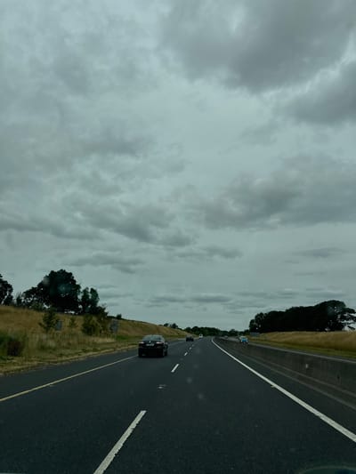 The image depicts a cloudy, overcast day on a highway in Hacklow, Ireland. The perspective is from inside a moving vehicle, looking forward along the road. The highway has multiple lanes, separated by white and yellow lines. Several cars are visible on the road, including a dark sedan in the foreground and a light blue car further ahead. The roadside features dry, grassy hills and dense trees along the right side of the highway, separated by a concrete barrier. The sky is filled with heavy, gray clouds, suggesting a possibility of rain. There are no discernible individuals in the scene, only the vehicles and the landscape.