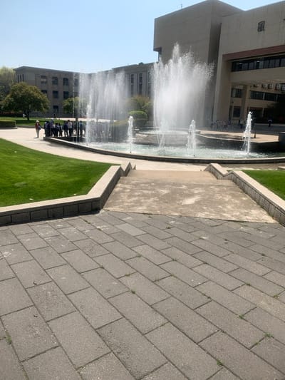 The image shows a fountain with several water jets spraying into a large circular basin in front of a building. There is a group of people standing near the edge of the basin. The area in front of the fountain is paved with gray bricks and there is a concrete ramp leading up to the fountain. The image appears to be taken from a street-level vantage point, looking down at the fountain from a slightly raised position. The image was taken in front of a large building, likely an academic building, located at Wuhan University (latitude: 30.516, longitude: 114.407). The weather is clear and sunny. The trees are green.