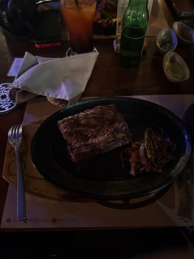The image depicts a table setting in what appears to be a restaurant. A black cast iron skillet is centrally positioned, containing a grilled steak and sautéed onions. A metal fork rests on the table next to the skillet. To the left of the skillet, there's a napkin, a green glass bottle, a cocktail glass filled with a reddish beverage, and a black wallet. In the upper left corner, there are three small glass bowls. The table appears to be wooden. A partial image of what looks like a placemat with the text "@otilia_mx" and social media icons indicates that the restaurant is likely "Otilia" in Tuxtla Gutiérrez, Mexico. No people are visible.