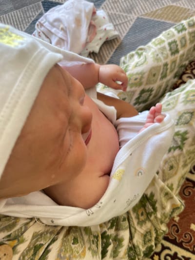 A close-up, high-angle shot shows a newborn baby's face and body, swaddled in white cloth with subtle colorful prints of rainbows and yellow shapes. A person's hand, with visible fingernails and skin texture, gently touches the baby's cheek, close to its mouth. The baby appears to be lying on a patterned surface, with textures suggesting fabric like a blanket or rug. The background shows a blurred pattern of a blue and gray geometric rug and a green and white floral fabric. The scene is indoors, with soft lighting. No specific location in Bujum, Kyrgyzstan is identifiable from the visual information provided.