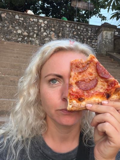 In Luang Prabang, Laos, a woman with blonde, curly hair holds a slice of pepperoni pizza up to her face, obscuring her left eye and nose. She is positioned in the foreground, looking towards the camera. Her right eye, which is visible, is blue-green. Behind her, a stone wall and a series of concrete steps are visible. Trees with green foliage overhang the wall in the background. The sky appears overcast. Two individuals are partially visible at the top of the steps, near the wall. One is wearing white clothing, and the other a green cap. The overall setting suggests an outdoor urban environment.