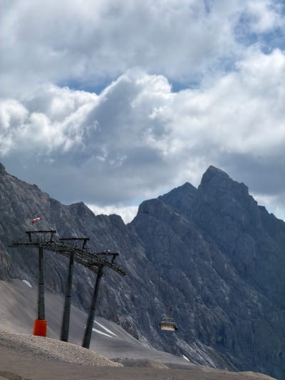 The image depicts a mountainous landscape with a ski lift in the foreground and jagged, rocky peaks in the background. The ski lift consists of several tall, grey towers with a complex system of wheels and cables. One of the towers has an orange cylindrical marker at its base. A single chairlift gondola is suspended from a cable in the mid-ground, to the right of the ski lift towers. A small red and white flag, possibly a windsock, is attached to the top of one of the ski lift towers on the left. The mountains are steep and covered in scree and sparse vegetation. The sky above is filled with dramatic, grey and white clouds, with patches of blue sky visible. The scene appears to be an alpine environment, likely at a ski resort. There are no people visible in the image. The location is the Zugspitze ski resort, which straddles the border between Germany and Austria, with the ski lift structures being on the Austrian side in the municipality of Wildermieming, Tyrol.