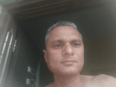A close-up, low-angle self-portrait of a middle-aged man with short, graying hair and light brown skin. He has a serious expression. He appears to be indoors, standing in front of a dark doorway or opening. No specific details visible indicate the location is Dhandhuka, India.