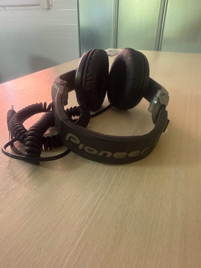 The image shows a pair of black Pioneer DJ headphones lying on a light-colored, wood-grain surface. The headphones are coiled and partially draped over themselves, with the earpads facing upwards. The headband is visible with the "Pioneer" logo printed in a lighter color. The textured surface suggests a desk or tabletop. In the background, to the left, is a wall with vertical panels, possibly in a room or a studio setting. The lighting appears to be artificial, casting a warm glow on the surface. There is no visible activity or people in the image. The overall scene is static and focused on the object. The location context is Britovo, Russia.