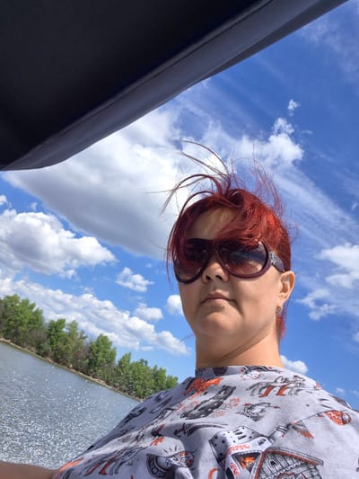 In a selfie taken outdoors, a woman with bright red hair, wearing large sunglasses, looks directly at the camera. Her hair is windswept. She is wearing a grey t-shirt with a pattern of animals and mythical creatures in black and orange. The background shows a bright blue sky with scattered white clouds. Below the sky, a body of water with choppy surface is visible, reflecting the light. Along the far bank of the water, a line of green trees is present. The scene appears to be a sunny day, possibly on a boat given the angle of the dark overhead structure in the upper left corner of the image, which suggests she is under some kind of canopy or awning. The location is Sady I Dachi, Russia.