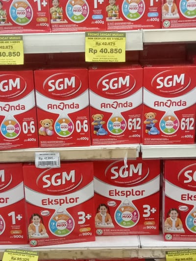 The image displays a retail store aisle featuring shelves stocked with various SGM brand powdered milk products. The specific location within Nganjuk, Indonesia, is not identifiable from the visual content.
The scene is dominated by three white, horizontally-aligned metal shelves filled with red and white cardboard boxes of SGM milk. The top shelf presents SGM Eksplor 1+ variants, primarily 400g boxes in "Madu" (Honey) and "Vanila" flavors, each adorned with an image of a smiling child. A prominent yellow promotional tag on this shelf advertises "PROMO SANGAT MURAH" (Very Cheap Promo) for SGM Eksplor 400g, displaying a reduced price of Rp 40.850 from Rp 42.875.
The middle shelf showcases SGM Ananda powdered milk in 400g boxes, specifically for ages 0-6 months and 6-12 months, featuring illustrations of teddy bears. A white price tag indicates a price of Rp 42.895 for SGM Ananda 400g.
The bottom shelf contains larger SGM Eksplor 1+ and 3+ variants, packaged in 900g red and white boxes, also depicting smiling children. A yellow promotional tag is partially visible at the bottom left, showing a price of Rp 85.325 for SGM Eksplor 900g.
All products are neatly arranged and uniformly stacked on the shelves. The environment suggests a brightly lit interior, likely a supermarket or large grocery store. No individuals are visible in the frame.