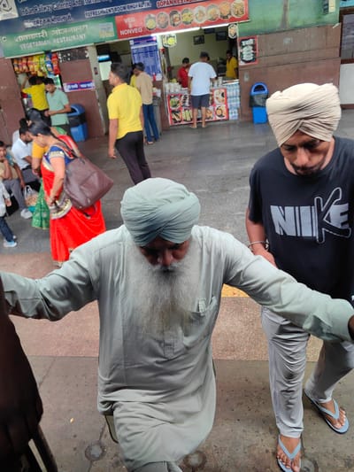 The image depicts a bustling scene at a railway station in Bhopal, India. In the foreground, two men, both wearing turbans, are walking. The man on the right wears a black Nike t-shirt and light grey pants, while the man on the left is dressed in a light grey kurta and pants. Behind them, several other people are visible, some queuing at a food stall with signage in Hindi, indicating "Vegetarian Restaurant" and "Veg & Non-Veg." The environment appears to be an outdoor area of the station, with tiled flooring and a weathered facade of buildings. The atmosphere suggests a busy commercial area within the railway station premises.