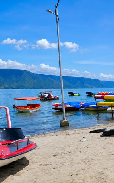 The image depicts a sunny day at the beach in Situngkir, Indonesia. The foreground features a sandy beach with two red and white boats resting on the shore, one with a black seat. In the shallow water, a pink structure is visible on the left, and a small orange boat with a canopy is anchored. Further out in the bright blue water, several colorful boats are floating, including a red and white motorboat with an outboard engine, and two yellow and green pedal boats. The background is dominated by a large, blue mountain range under a clear blue sky with scattered white clouds. A tall, grey lamppost stands in the middle ground, casting a shadow on the sand. The overall scene conveys a sense of a peaceful and recreational lakeside environment.