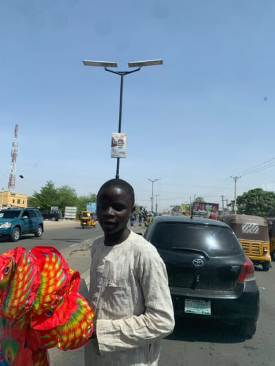 The image depicts a daytime street scene in Hotoro, Nigeria, under a clear, light blue sky. In the foreground, a young Black male, appearing to be in his late teens or early twenties, stands slightly to the left of the frame, facing partially towards the camera. He wears a light-colored, long-sleeved, collared garment with faint vertical stripes. He holds a bundle of brightly colored objects, possibly folded textiles or lanterns, displaying radial patterns in red, orange, yellow, green, and blue.
Immediately behind him and to his right is a black Toyota Yaris hatchback with a visible Nigerian license plate from Kano state, displaying "DAI 77C0". Further into the midground, an asphalt road features moderate vehicle traffic, including a blue SUV on the left, a yellow motorized rickshaw (Keke Napep) on the right, and other indistinct vehicles.
A tall black street light pole with two horizontal solar-powered LED luminaires at its apex stands prominently in the center of the image, behind the primary subject. A rectangular banner depicting a man in a red cap with accompanying text is affixed to this pole. In the background, on the far left, a red and white cellular tower is visible. Scattered low-rise buildings and sparse green vegetation line both sides of the road. Multiple indistinct pedestrians are visible along the road and sidewalks. The scene is brightly lit by natural sunlight, indicating midday conditions.