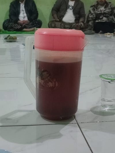 The image shows a red liquid in a clear pitcher with a pink lid and a clear handle. There is a clear glass with a green lid and a white liquid to the right of the pitcher. The background shows three men sitting on a light-colored tiled floor. They are wearing various clothing, and the two visible men have their arms crossed. The men appear to be of an older age. The setting is a tiled room in Kecamatan Pandaan, Indonesia, with coordinates -7.668, 112.724. The tiled floor is made of rectangular white and gray tiles. The pitcher is sitting on the tiles in the center of the image. The glass is to the right of the pitcher. The men are in the background, and their faces are not visible. The light is coming from the left side of the image.