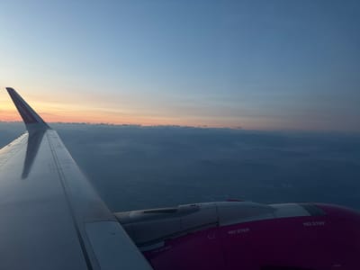 The image is an aerial view from an airplane window, likely captured during sunrise or sunset, given the soft, gradient sky with hues of blue, pink, and orange. In the foreground, a portion of the airplane's wing is visible, extending from the left edge of the frame. The wing is metallic and appears to be white or silver, reflecting the ambient light. A portion of the airplane's engine housing, painted in a vibrant magenta color, is visible in the lower right quadrant of the image. Text, including "NO STEP" and "wizzair.com," is visible on the wing and engine components.

Below the wing and engine, a landscape of rolling hills or mountains is visible, shrouded in a light mist or haze, creating a sense of depth and distance. Some faint signs of human habitation, such as scattered lights or structures, can be discerned within the darkened landscape, suggesting a rural or semi-urban area. The scene depicts a moment of flight over a serene, atmospheric landscape.

Regarding the location in the city of Caravaggio, Italy: While the image depicts a landscape that could be consistent with the geographical features of the Lombardy region where Caravaggio is located, there is no specific visual information within the image that definitively places it in Caravaggio itself. The landscape is generic enough to be many mountainous or hilly regions, and the aerial perspective does not offer identifiable landmarks of Caravaggio.