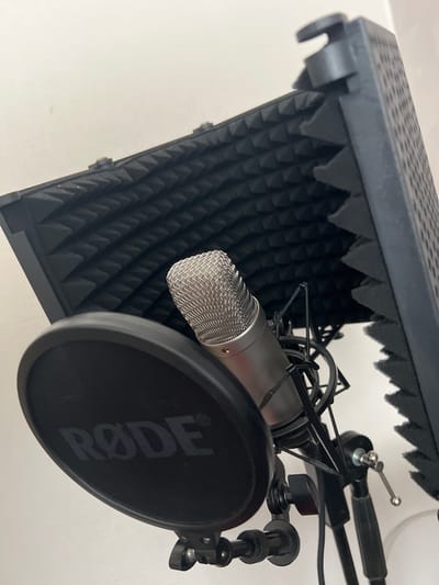 This media file shows a close-up view of a recording setup. The main objects are a silver microphone, a black pop filter with the "RØDE" logo visible, and a black sound-absorbing shield with a triangular foam pattern. The microphone is mounted on a stand, which is partially visible. The setting appears to be indoors, with a plain white background. The lighting suggests it could be daytime, but there are no external cues to confirm this. The arrangement of the equipment indicates it is set up for audio recording, possibly for voiceovers, podcasts, or music. There are no people or animals in the frame. The overall impression is one of a functional, professional audio recording environment. There are no specific location cues that would strongly suggest Ventspils, Latvia, beyond the provided context.