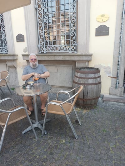 An elderly man with a white beard and glasses sits at a round metal table outside a building in Casalmaggiore, Italy. He wears a grey t-shirt and dark shorts, with brown sandals. In front of him on the table is a glass of orange liquid, a small bowl of chips, and an ashtray with a mask. The man is seated in a wicker chair with a metal frame. To his left is another empty chair of the same design. Behind the table and chairs, a large wooden barrel is visible, leaning against the building. The building has cream-colored walls and a large window with intricate metal bars. Two plaques are affixed to the building's wall near the window. The ground is paved with dark cobblestones, with patches of grass growing between them.