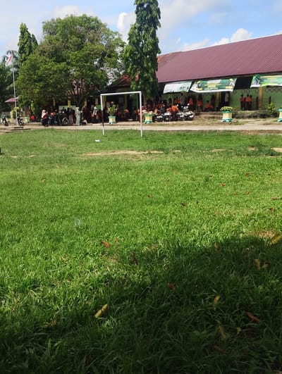 The image captures an outdoor scene, likely a schoolyard in Pantonrayeuk Satu, Indonesia. In the foreground, a vast expanse of well-maintained green grass is visible, with scattered brown leaves and a few small pieces of litter. A shadow from an unseen object falls across the right side of the grass.
In the midground, a white goal frame stands on a paved area. Behind the goal, a building with a dark red roof and green walls is present. Several banners with green backgrounds and white text are displayed along the building's facade. A group of people, appearing to be students and possibly staff, are gathered in front of the building, some sitting on steps and others standing. Many of them are wearing orange attire. Several motorcycles are parked in the vicinity.
To the left of the building, a tall, slender tree with dark green foliage stands. Further left, a flagpole with the Indonesian flag is visible, accompanied by another large tree with dense green foliage. The sky above is blue with scattered white clouds. The overall impression is of a school event or gathering taking place on a sunny day.