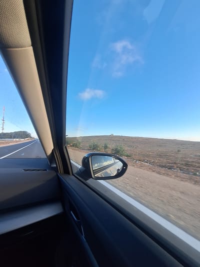 The image captures a view from the passenger side of a car, looking outwards and slightly upwards. The scene is dominated by a vast, bright blue sky with a few wispy white clouds. To the right, a dry, brownish landscape stretches to the horizon, suggesting an arid or semi-arid region. A road is visible, bordered by this terrain, and a portion of the car's interior, including the door frame and part of the dashboard, frames the view. A side mirror is attached to the car door, reflecting the sky and the passing landscape. The car appears to be in motion on a road, indicated by the blurred motion of the scenery and the perspective. The time of day appears to be daytime, likely late morning or early afternoon, given the bright sunlight and clear sky. The overall impression is one of travel through an open, possibly rural or undeveloped area. There is no discernible text within the image.