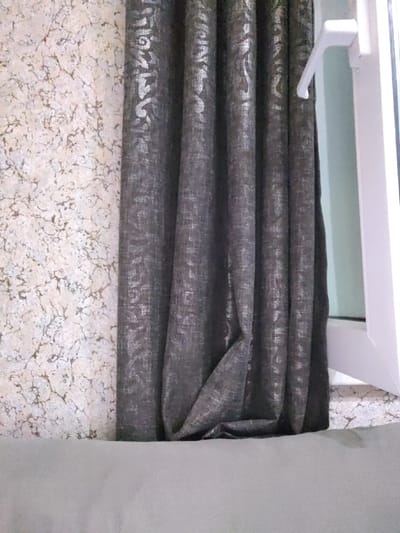 This media file captures a close-up view of a window area within a room. The primary subjects are dark, patterned curtains partially obscuring a window with a white frame and handle. To the left of the curtains, a textured wallpaper with a light, mottled pattern is visible. In the foreground, a pale gray, upholstered surface, likely a sofa or bed, occupies the bottom portion of the frame.

The scene is static, with no discernible activity taking place. Notable details include the reflective pattern on the dark curtains, which appears to be a silver or metallic floral design. The window handle is in the open position, suggesting the window might be ajar. The lighting appears to be natural, coming from the window, and there are no cues about the time of day or weather outside. The overall impression is of an indoor domestic setting, with elements of interior decoration. There is no visible text in the image.