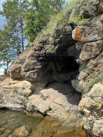 The image shows a rocky cliff face with a dark cave opening near the bottom right. The cliff is made of large, rough-textured rocks with patches of orange lichen and green vegetation. Trees, including pine trees with needles and a deciduous tree with green leaves, grow at the top of the cliff. The base of the cliff meets a body of clear, shallow water with submerged rocks visible. The sky is partly cloudy. There are no people or animals visible in the image. The scene appears to be a natural outdoor environment.