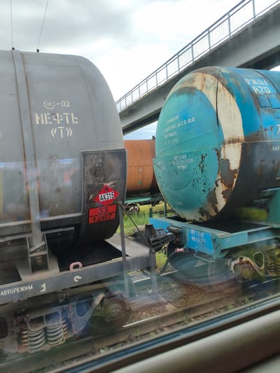 The image depicts several railway tank cars parked on tracks under a pedestrian overpass in Buzuluk, Russia. The foreground features the side of a weathered grey tank car with Cyrillic lettering that reads "НЕФТЬ" ("NEFT") and "«Т»". A red diamond-shaped hazard symbol with "AK315" and the numbers "33" and "1267" is affixed to its side. To the right, a large, rusted, turquoise tank car is visible, also bearing Cyrillic inscriptions and a crack running down its middle. The background shows a concrete overpass with a metal railing, and beyond that, a cloudy sky. Another orange tank car is partially visible between the grey and turquoise ones. The overall scene suggests an industrial or transportation hub within the city.