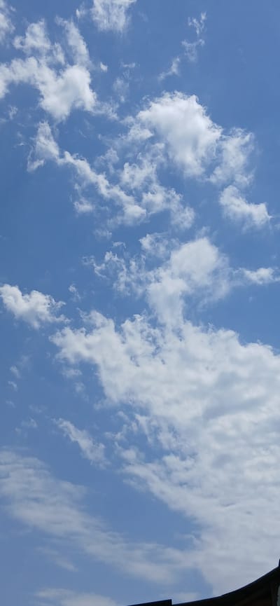 The image depicts a sky filled with scattered white clouds against a clear blue backdrop. The clouds vary in size and density, with some appearing fluffy and cumulus-like, while others are thinner and more wispy. The sunlight illuminates the clouds, giving them a bright, ethereal quality. On the right side of the frame, a dark, curved object partially obscures the sky, suggesting the presence of a structure or an overhang. The scene appears to be an outdoor setting on a bright, clear day, likely during daylight hours due to the illumination of the clouds. There are no people or discernable objects within the sky itself. The composition and the vast expanse of the sky evoke a sense of openness and tranquility. The image does not contain any visible text. The location context provided is Kuchtumgali, Uzbekistan.