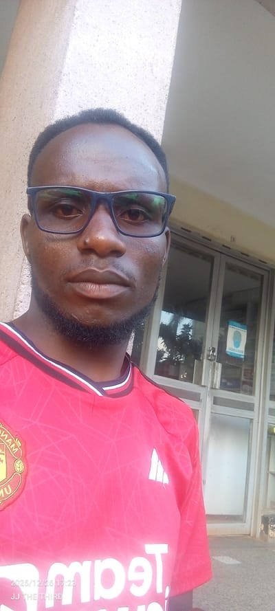 The image displays an adult dark-skinned male, positioned outdoors in Kisii Central ward, Kenya. He is wearing dark-rimmed glasses and has a short beard. His attire is a red short-sleeved Manchester United football club jersey, featuring the club crest on the left chest and an Adidas logo on the right. Reversed white lettering, partially visible across the chest, reads "TeamViewer". The subject faces the camera directly. The background consists of a light brown textured wall to the left and a building entrance with multiple glass panels to the right. A blue and white sign with a stylized masked face and reversed text "WEAR A MASK" is affixed to the right glass panel of the door. Reflections of green foliage are visible through the glass. A timestamp "2025/12/26 17:23" and text "JJ THE THIRD" are present in the lower-left corner of the image.