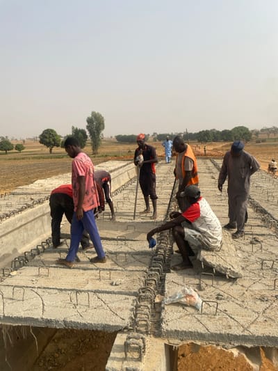 The scene depicts a construction site in Wuciciri, Nigeria, under a hazy, pale sky. Approximately ten adult males are present on and around a partially constructed concrete structure. In the foreground, six men are directly on the structure, which displays a grid of exposed rebar stirrups embedded in fresh concrete, indicative of a reinforced concrete slab or bridge deck under construction.
A man in a pink t-shirt and blue pants walks across the concrete structure. Two men are bent over, appearing to work on the rebar or concrete. A man wearing an orange safety vest stands holding a long tool, while another man in dark grey coveralls stands to his right. A fifth man, dressed in a red and white t-shirt and shorts, squats on the concrete, holding a blue cloth. A white plastic bag rests on the concrete near the squatting man.
The background shows a flat, arid landscape with scattered trees and dry, brownish fields. A dirt path extends into the distance on the right, where additional figures and a motorcycle are visible.