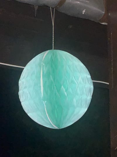 A turquoise spherical paper lantern hangs against a dark, textured background. The lantern has a diamond-like pattern formed by creases in the paper and a white band around its middle. A thin white string, likely for hanging, is attached to the top of the lantern and extends upwards out of frame. Another string is attached to the left side of the lantern and extends horizontally to the left, connecting to a dark, cylindrical object wrapped in plastic. The object appears to be a bundle of something. The overall scene is dimly lit, suggesting it might be indoors or nighttime. The location is identified as Gradets, Bulgaria. No people are visible, and no text is present in the image. The focus is on the lantern and the attached object, with the dark background providing contrast.