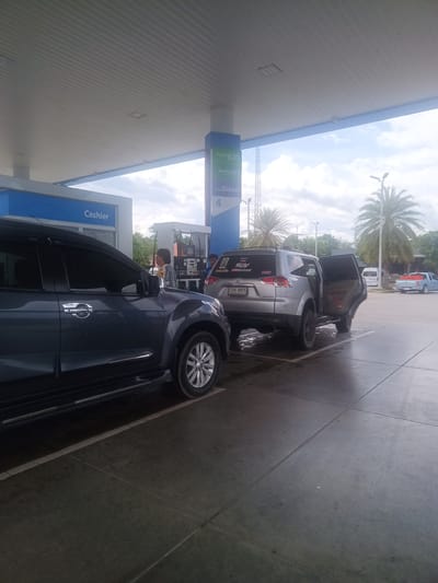 This is an outdoor image taken at a gas station in Tha Ruea, Thailand. The scene features two vehicles. In the foreground, a dark grey Isuzu MU-X is parked with a partial view of its side. Behind it, a silver Mitsubishi Pajero Sport SUV is being filled with fuel, as indicated by a person standing near the gas pump. The SUV's back door is open.

The gas station's infrastructure includes a "Cashier" building on the left and a gas pump island with a sign indicating different fuel types (Gasohol E20, Gasohol 91, and Diesel) and the pump number "4."

In the background, beyond the gas station, is a palm tree and another light blue pick-up vehicle is parked farther away, and a white van is located between the two vehicles. The sky is overcast with light grey clouds.