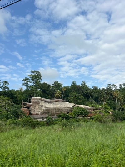 The image depicts a concrete structure surrounded by lush vegetation, likely in Walahanduwa, Sri Lanka. The building appears to be in a state of disrepair or under construction, as scaffolding is visible around its exterior. The surrounding environment features dense greenery, including trees, shrubs, and tall grass, which is commonly found in tropical climates. The sky is partially cloudy, suggesting a humid day. The absence of people and specific activities allows a focus on the structural and environmental aspects of the scene.