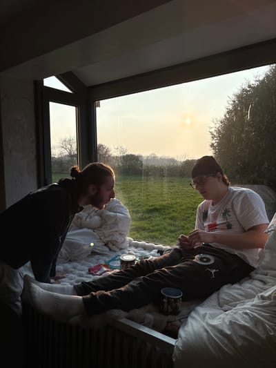 The image displays an indoor scene in Bû, France, featuring two males positioned near a large window that overlooks a grassy field with trees under a low sun. The male on the left, with reddish-brown hair tied in a bun and a beard, wears a dark hoodie and light pants, leaning forward towards the other individual. The male on the right is seated on white, padded bedding, wearing a black beanie, eyeglasses, a white t-shirt with a palm tree graphic, dark sweatpants with a circular logo, and white socks. He is actively manipulating small objects with his hands. Various items, including small containers, a red packet, and a dark mug, are present on the bedding between the two subjects. The setting sun is visible through the window, casting a golden light across the scene and the outdoor landscape.