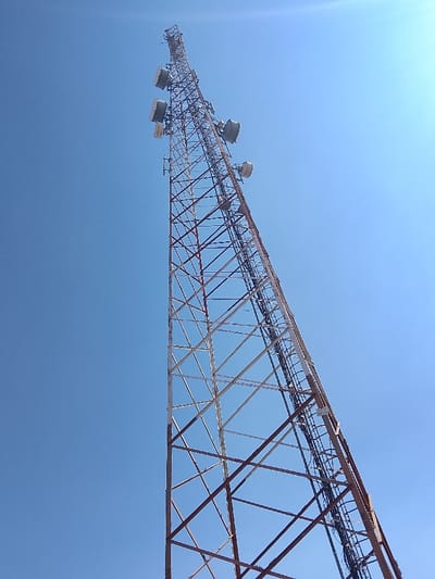 A telecommunications lattice tower is observed in Vodni, Nigeria, photographed from a low-angle perspective against an unobstructed blue sky. The tower, constructed from metal trusses, tapers upwards, displaying a surface with sections of reddish-brown and lighter-toned material. Multiple rectangular panel antennas are mounted at various elevations on the tower's upper structure, complemented by at least one smaller, circular dish antenna. Black electrical cables are secured in vertical bundles along one structural leg. The ambient lighting suggests clear daylight conditions, with no visible personnel or ground-level infrastructure.