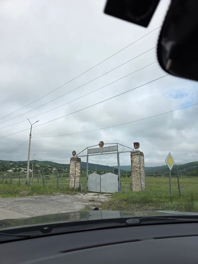 The image shows an overcast sky above a rural landscape. In the foreground, the dashboard of a vehicle is visible, suggesting the photo was taken from inside a car. A gray road leads towards a large, imposing gate structure. The gate consists of two weathered brick pillars topped with decorative metal elements, supporting a metal archway. On the archway, there are circular emblems with intricate designs. Behind the gate, a pair of closed metal gates are visible.
To the left of the gate, a tall, slender lamppost stands, with a smaller lamppost further in the background. In the distance, a village with houses and fences is nestled on a rolling green hillside. Power lines stretch across the sky, supported by poles. To the right of the gate, a yellow diamond-shaped road sign is positioned. The overall environment appears to be the outskirts of the city of Теплоозёрск, Russia, characterized by a mixture of natural landscape, rural infrastructure, and a cloudy atmosphere. No people are visible in the frame.