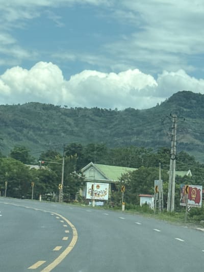 This is a photograph taken from a moving vehicle, looking out at a road bordered by lush greenery and a towering, forested mountain. The sky is a bright blue with large, fluffy white clouds. The road curves to the right, with a yellow lane marker and white dashed lines indicating it is a paved road. Along the side of the road, there are several objects, including utility poles with wires, streetlights, and various signs.
One sign, prominently displayed on a billboard, features a red and white design with what appears to be an image of a person. Another smaller sign below it has a similar color scheme. Further down the road, another billboard shows colorful graphics, possibly advertisements. A light green building with a peaked roof is visible amongst the trees, suggesting a roadside establishment or residence.
The scene suggests a bright, clear day, likely in the late morning or early afternoon given the lighting. The abundance of greenery and the presence of the mountain imply a rural or semi-rural location, consistent with the provided context of Lao Bảo Commune, Vietnam. The overall impression is one of a scenic drive through a tropical or subtropical landscape. There is no indication of people in the immediate frame, but the presence of a road and signs suggests human activity in the area.