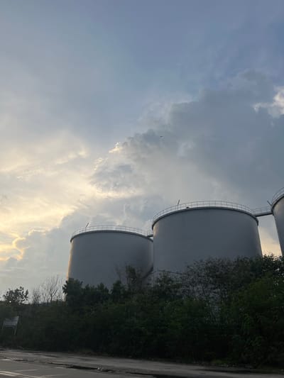 The image depicts an industrial scene featuring multiple large, cylindrical storage tanks, likely part of a depot or refinery complex in Dumai City, Indonesia. The tanks are painted a light gray and are topped with railings and access walkways. They are partially obscured by lush green foliage and scattered trees in the foreground. The sky above is dramatic, with a mix of dark, heavy clouds and patches of bright, illuminated clouds, suggesting either an approaching storm or a sunset. A single bird is visible in the sky. The foreground shows a paved area, possibly a road, with some puddles, indicating recent rain. The overall impression is one of industrial infrastructure set against a dynamic natural environment.