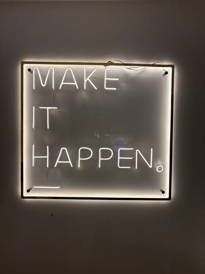 A square neon sign with the words "MAKE IT HAPPEN" vertically arranged hangs on a light-colored wall. The sign has a metallic frame with illuminated edges, and the lettering is in a bright white neon. The words are oriented in two columns, with "MAKE IT" on the left and "HAPPEN" on the right. The background of the sign is a slightly reflective white or light gray. The wall is plain with a subtle texture. The location is unknown.