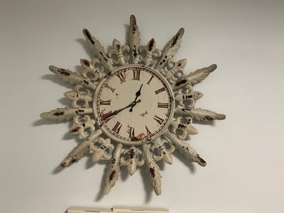 The image shows a close-up, eye-level, indoor shot of a decorative wall clock. The clock has a cream-colored, distressed finish with visible brown marks, giving it a vintage appearance. It features a circular face with Roman numerals representing the hours. The hour hand points to approximately 10, and the minute hand points to approximately 6, indicating the time is roughly 10:30. The clock is surrounded by an ornate, sunburst-like frame made of sculpted elements resembling feathers and fleur-de-lis. These decorative elements are also cream-colored and show the same distressed finish. The clock is mounted on a plain white wall, which provides a clean backdrop and highlights the clock's intricate design. The lighting appears to be ambient indoor light, casting a subtle shadow from the clock onto the wall, suggesting it is not directly lit from the front. There is no visible text in the image. The setting is likely a home or a public space where decorative clocks are displayed.