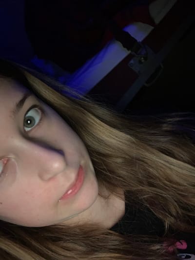 A close-up, angled selfie shows a woman with long, light brown hair. Her face is pale, with a noticeable nose and slightly parted lips. Her eyes are wide, with a light green iris visible in one. The background is dark and blurred, suggesting a low-light environment. A faint blue glow illuminates part of the background, highlighting a metallic structure with a handle and straps. The overall image is slightly distorted due to the angle.