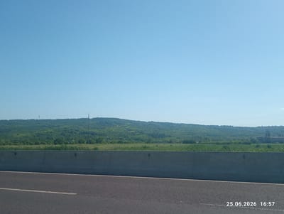 The image shows a rural landscape under a clear blue sky. In the foreground, a portion of an asphalt road with a white lane marker is visible, leading towards a concrete barrier. Beyond the barrier, a dense, green hillside covered in trees and vegetation stretches across the horizon. There are no people or animals in the frame. The time and date stamp "25.06.2026 16:57" is visible in the lower right corner. The location is identified as Cojușna, Moldova.