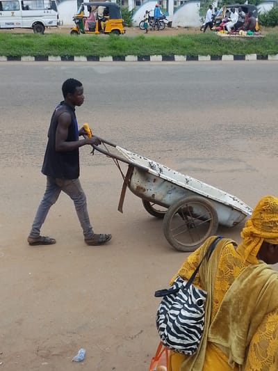 This media file depicts an outdoor scene in Oja Oba, Oko Erin, Nigeria. The primary subject is a man pushing a two-wheeled cart along a paved road. He is wearing a dark sleeveless shirt, light blue jeans, and dark sandals. In his right hand, he holds a yellow object that appears to be a corn cob. The cart is made of metal and shows signs of wear and tear.
In the foreground, to the right of the cart, a woman wearing a yellow headscarf and a yellow patterned outfit is visible. She is carrying a black and white zebra-striped bag.
In the background, various vehicles are present, including a white van on the left and a yellow three-wheeled motorized vehicle with passengers. Further in the background, more people and vehicles are present, suggesting a busy street or marketplace. The setting appears to be a roadside with some vegetation and a black and white curb. The overall lighting suggests it is daytime, with no specific indicators of the weather. There is no visible text in the image. The activity appears to be transportation of goods or materials via the cart, common in such locations.