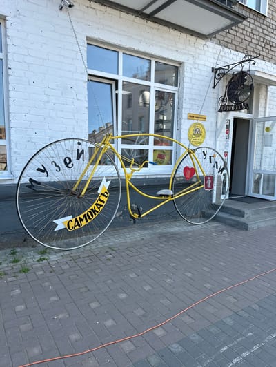 A large, yellow, novelty bicycle sculpture is displayed outside a building in Nikul'skoe (Neverkovskii S/O), Russia. The bicycle has extremely oversized wheels, with the front wheel significantly larger than the rear. Black lettering is painted on the spokes of the front wheel, reading "МУЗЕЙ" (MUSEUM), and on a yellow banner attached to the wheel, reading "САМОКАТЬ" (ROLL). Black lettering on the rear wheel reads "УГЛИ" (ANGLES). A red heart is attached to the frame near the rear wheel. The building features white brick walls and large windows. A sign with the text "Васи Самкатова" and operating hours is visible above the entrance. A security camera is mounted on the wall above the bicycle. The bicycle is suspended by chains. The ground in front of the building is paved with brick.