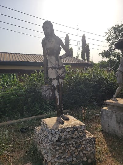 The scene depicts an outdoor public space in San Martín, Colombia, featuring a weathered statue of a standing female figure. The statue, appearing to be cast metal with significant paint peeling revealing a lighter underlayer, faces slightly left with her head tilted downwards. Her left arm is bent, hand on her hip, while her right arm holds a rectangular bucket or basket. She stands on a two-tiered base: a lower section composed of irregular, light-colored stones set in mortar, topped by a concrete slab. To the right, partially visible, is another statue on a similar concrete base, showing a lower torso and legs in motion. Dense green foliage and sparse dry grass cover the ground around the statues. In the background, a building with a light-colored corrugated metal roof is visible behind the foliage. Overhead, multiple power lines stretch across a bright, clear sky. Tall, slender coniferous trees and a distant lattice-structured communication tower are also discernible in the background. The lighting suggests bright daylight.