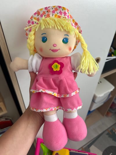 A stuffed doll with blonde yarn hair, blue eyes, a pink mouth, and rosy cheeks is being held by a person's hand. The doll is wearing a floral print bonnet, a pink dress with a yellow and pink flower applique, and pink shoes. The background appears to be an indoor setting with white walls and shelves filled with various objects, suggesting a domestic or retail environment. The location is not specified within Belfast City, United Kingdom, as the image provides no contextual clues for this.