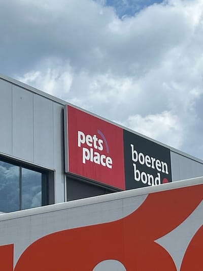 The image shows a building in Ede, Netherlands, featuring the logos of "Pets Place" and "Boerenbond" on its exterior. The building is a modern, angular structure with a silver-gray facade. The "Pets Place" logo is on a red background, while "Boerenbond" appears on a black background. A partial view of a large orange graphic is visible on the right side of the frame. A window reflects the sky. The background shows a partly cloudy sky.