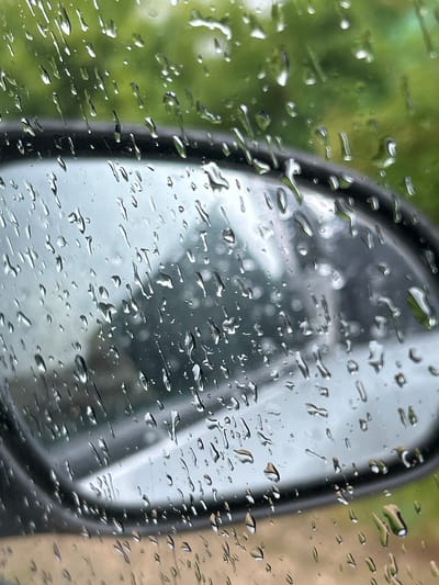 The image depicts a close-up view of a car's side mirror covered in raindrops. The mirror reflects a blurred outdoor scene, likely featuring trees and possibly other vehicles or structures, with a predominantly green and grey color palette. The foreground is dominated by numerous water droplets of varying sizes adhering to the glass surface, some appearing elongated as if having run down, while others are more spherical. The background shows a soft, out-of-focus green foliage. The car's dark grey mirror casing is visible along the bottom and left edges of the image. The overall impression is one of a rainy day. The location is specified as Kurinskaya, Russia.