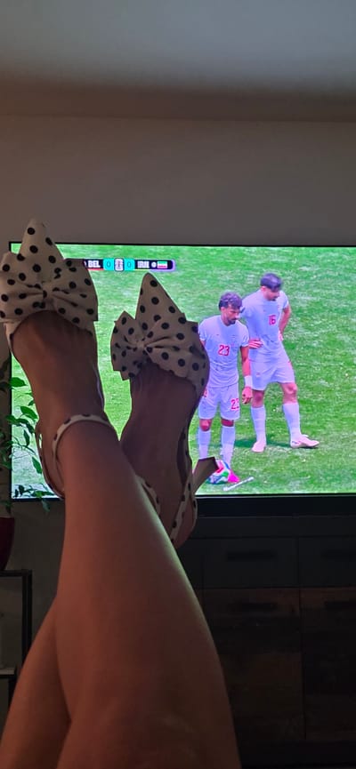 The image shows a person's legs, adorned with polka-dotted high heels, raised in front of a television screen. The television displays a soccer match between Belgium (BEL) and Iran (IRN), with the score currently 0-0. Two male soccer players in white jerseys are visible on the screen, one wearing the number 23 and the other number 16. The setting appears to be a living room, with a dark cabinet visible below the television. The scene is dimly lit, with the primary light source coming from the television. The image does not contain any information about the city of Lieli, Switzerland.
