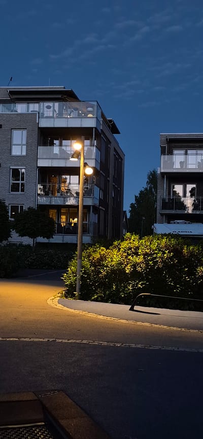 The image depicts an outdoor scene at dusk, featuring modern apartment buildings and a street lamp. The sky is a deep blue with sparse clouds. In the foreground, a paved road curves to the left, illuminated by a warm yellow glow from a street lamp. The lamp post is wooden and has two lights. To the right of the street lamp, a dense hedge is brightly lit from the side, creating dramatic shadows.

Behind the hedge and street lamp, two multi-story apartment buildings are visible. The building on the left is constructed with grey brick and features multiple balconies with glass railings. Lights are on inside some of the apartments, casting warm reflections. The building on the right also has balconies with glass railings and appears to be made of darker materials. A white trailer with "ADRIA" branding is partially visible in front of the building on the right. Trees and foliage are present around the buildings.

The image is static and there are no people or animals visible. The overall impression is one of a quiet, residential area at the end of the day. The specific location in Lillehammer, Norway, is not evident from the visual cues alone.